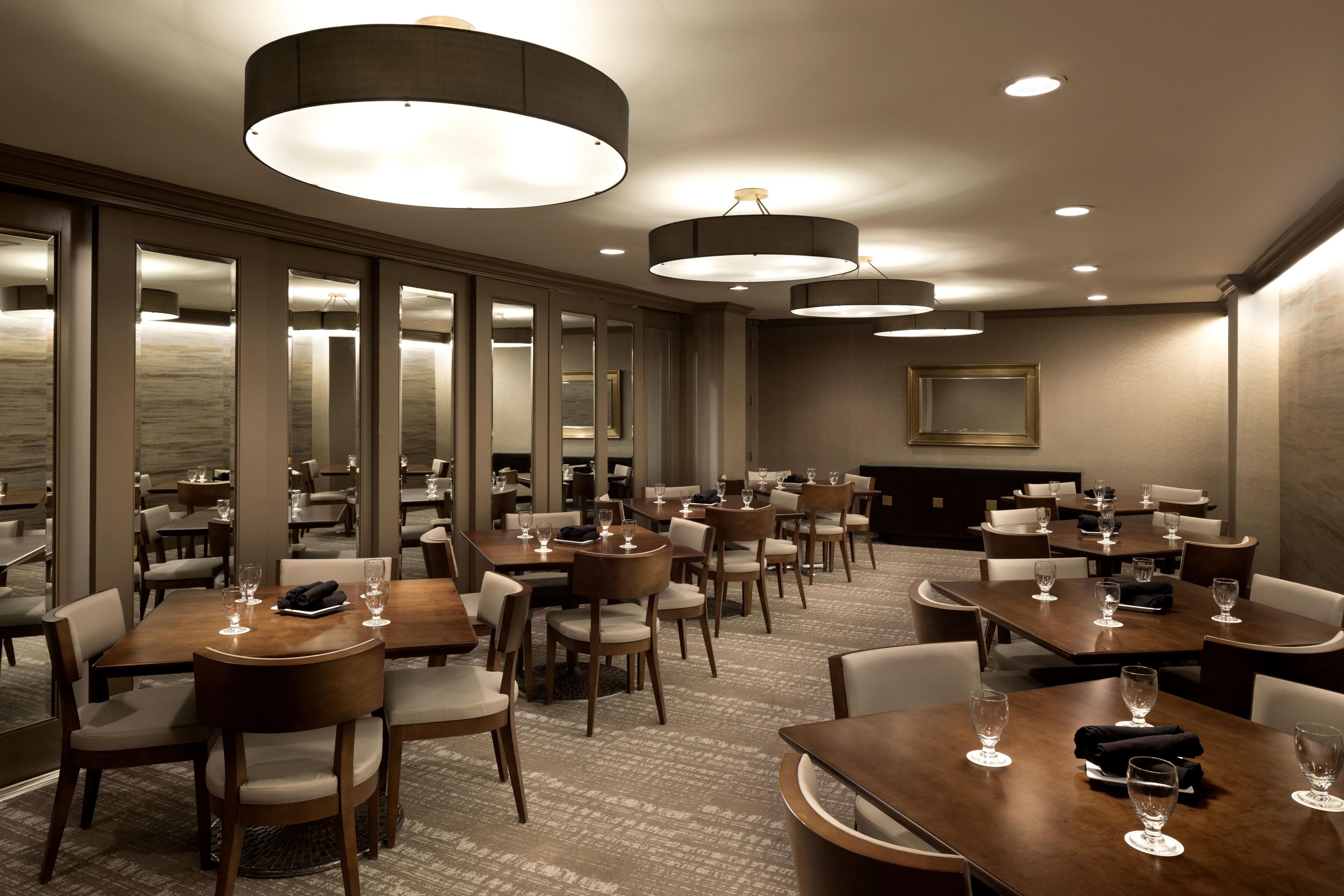 A hotel dining area with multiple tables and chairs, overhead lighting, and a large mirror on the wall.