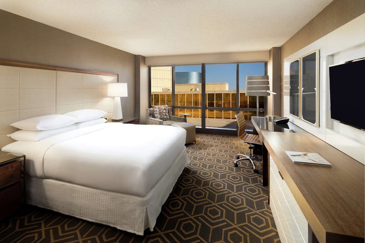 A hotel room with a large bed, a desk, a chair, a TV, and a balcony with a view of a building.