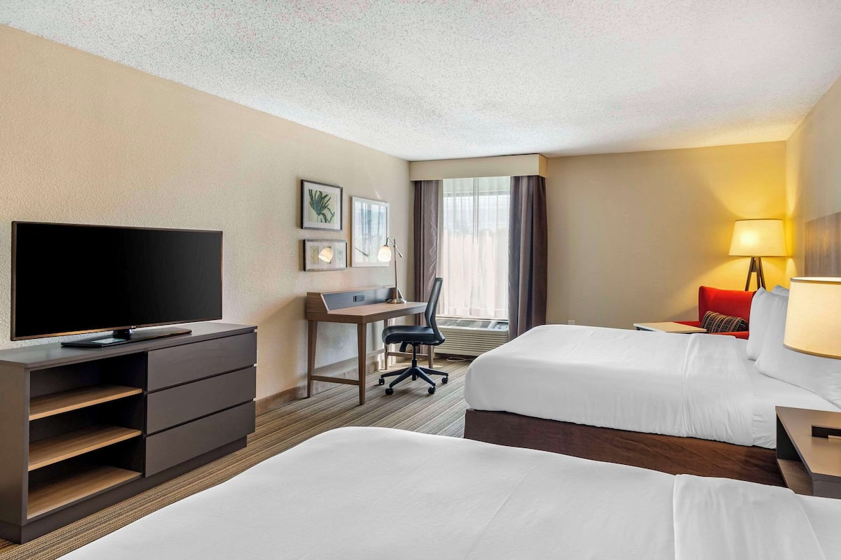 A hotel room with two beds, a desk with a chair, a flat-screen TV, a lamp, and a window with curtains.