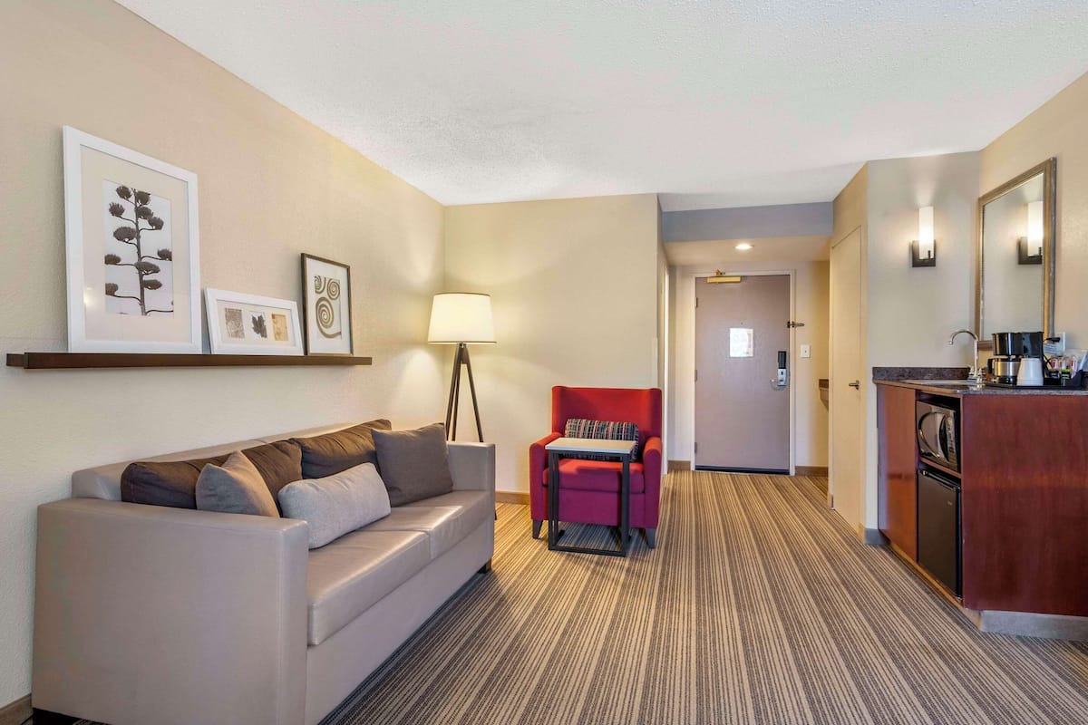 A hotel room with a sofa, a red armchair, a lamp, a kitchen area, and framed artwork on the wall.