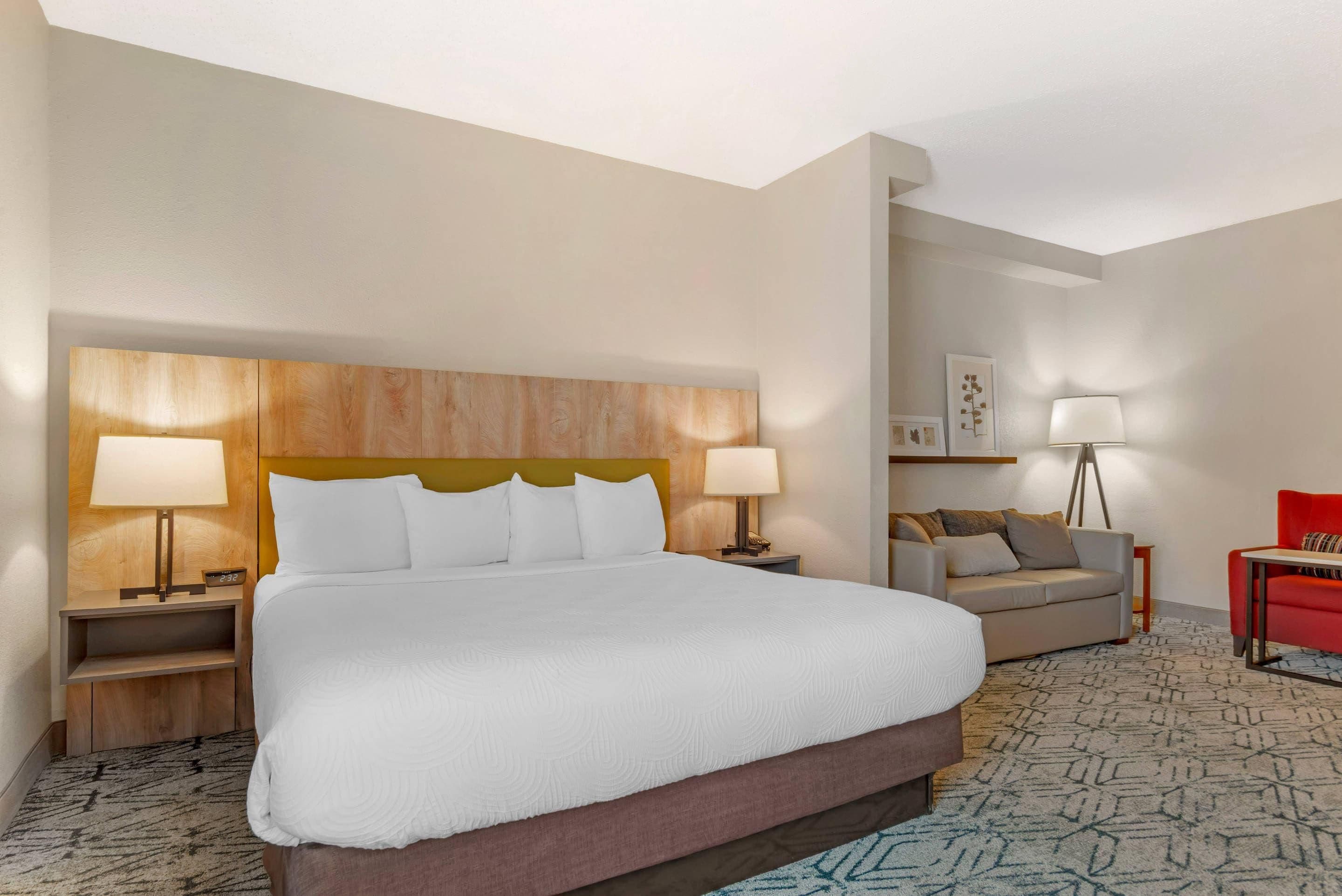 A hotel room with a large bed, two bedside tables with lamps, a sofa, a red chair, and a floor lamp.