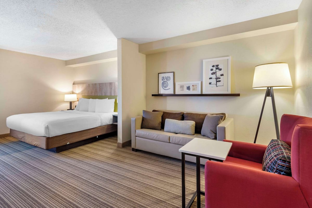 A hotel room with a bed, a sofa, a red armchair, a small table, and wall-mounted artwork.