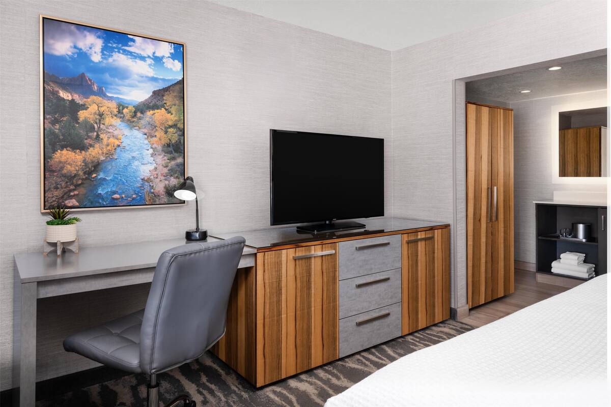 A modern hotel room with a wooden desk, a flat-screen TV, a grey office chair, a bed with white bedding, and a large window with a mountain view.