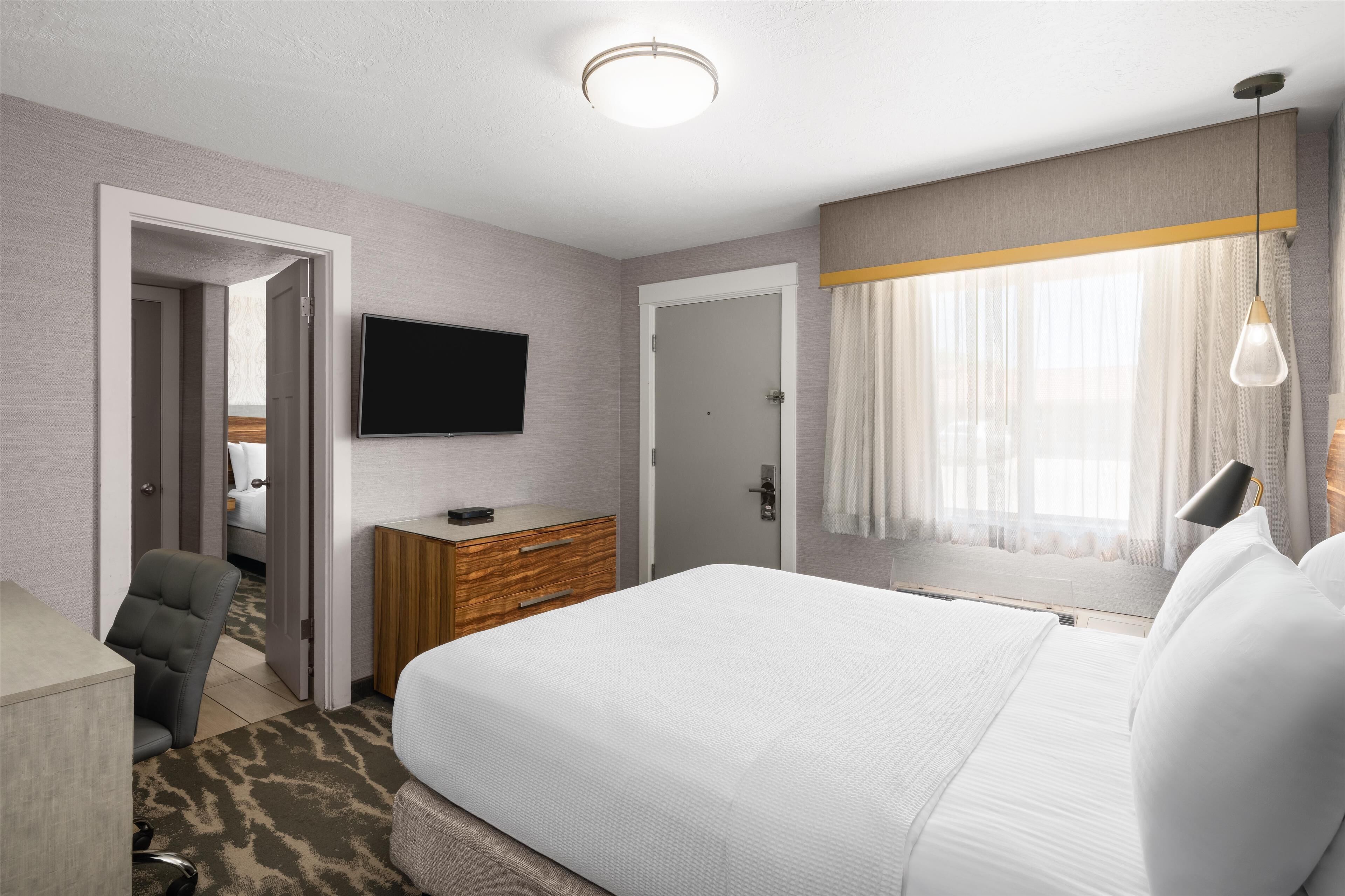 A hotel room with a large bed, a flat-screen TV mounted on the wall, a wooden dresser, a chair, and a window with sheer curtains.
