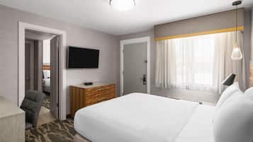 A hotel room with a large bed, a flat-screen TV mounted on the wall, a wooden dresser, a chair, and a window with sheer curtains.