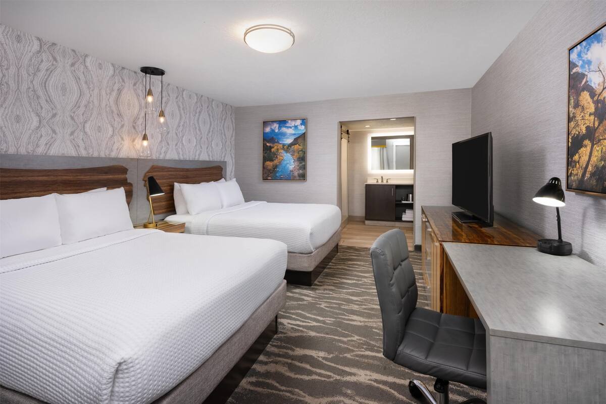 A hotel room with two beds, a desk with a TV, a chair, a painting, and a bathroom visible through an open door.