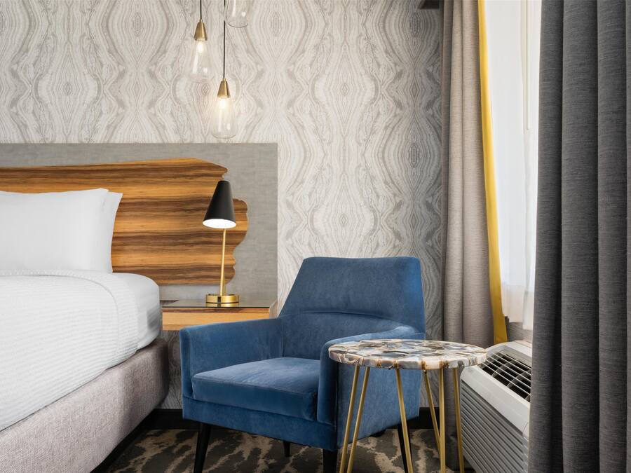A hotel room with a bed, a blue armchair, a small round table, a lamp, and a radiator.