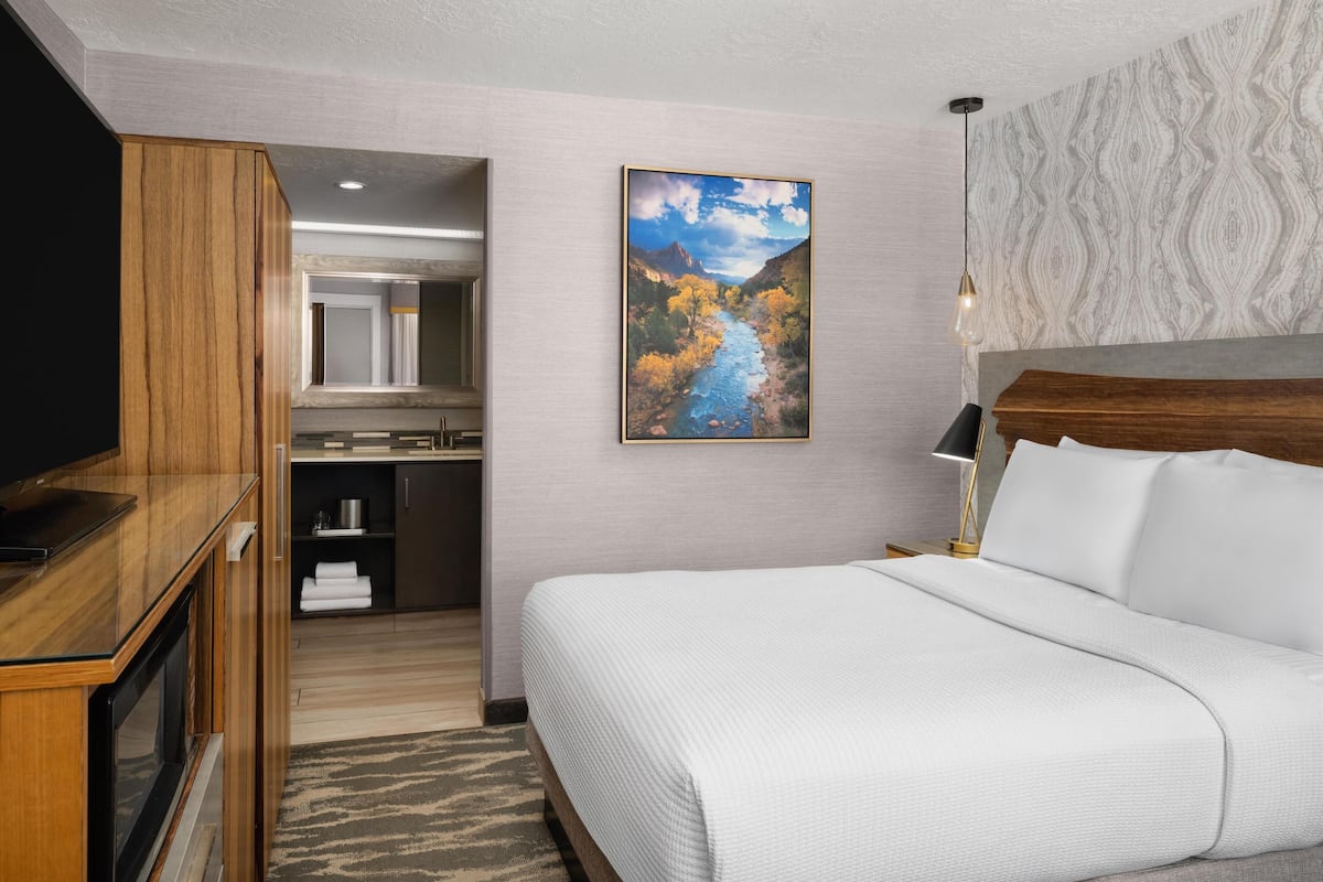 A hotel room with a bed, a TV, a painting of a river, and a bathroom visible through an open door.