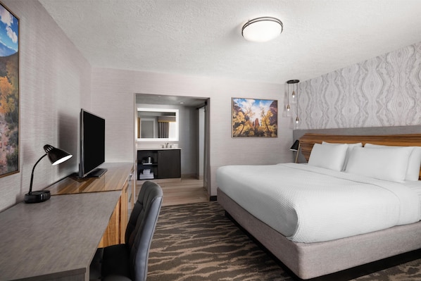 A hotel room with a bed, a desk with a TV, a desk lamp, a chair, a bathroom, and a painting on the wall.