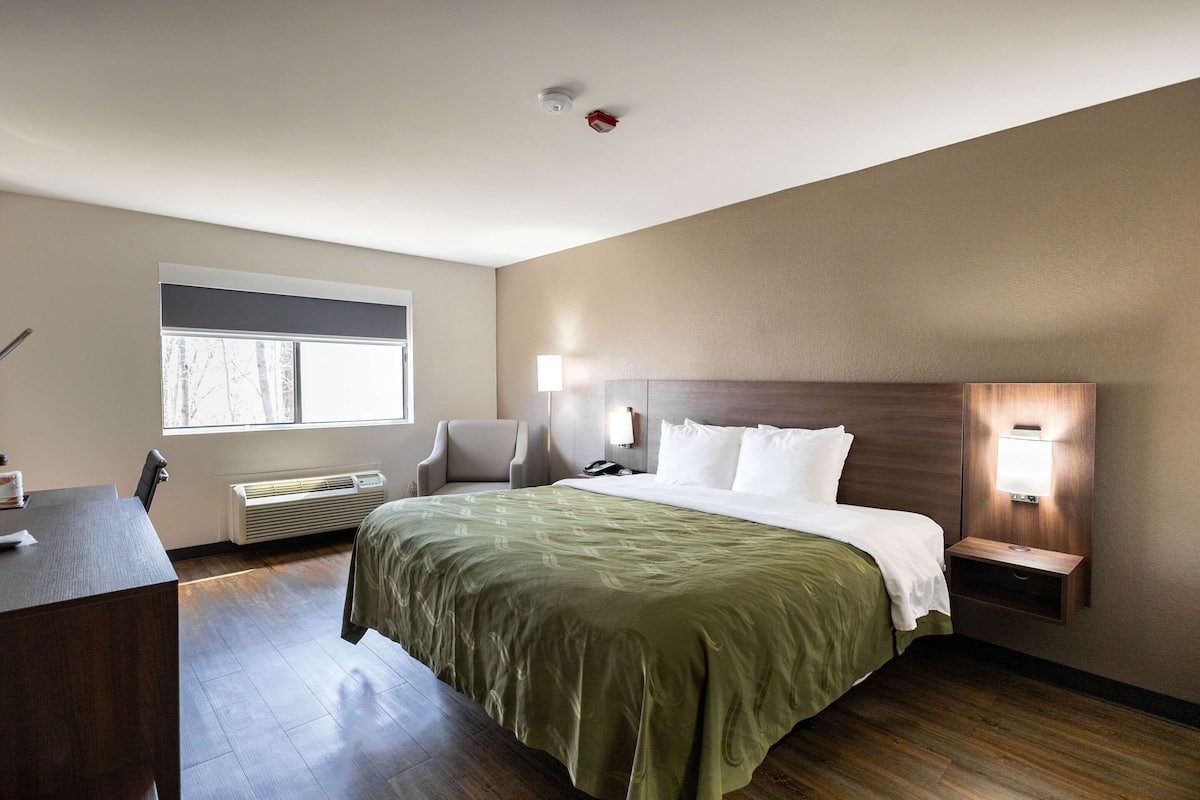 A hotel room with a bed, bedside table, lamp, chair, and a window.
