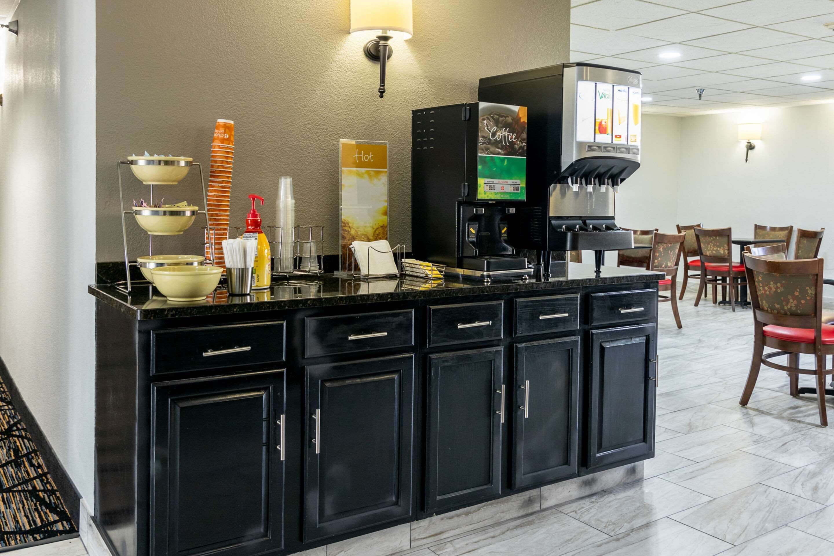 A hotel breakfast area with a self-serve coffee station, a stack of orange cups, and a sign indicating hot beverages.