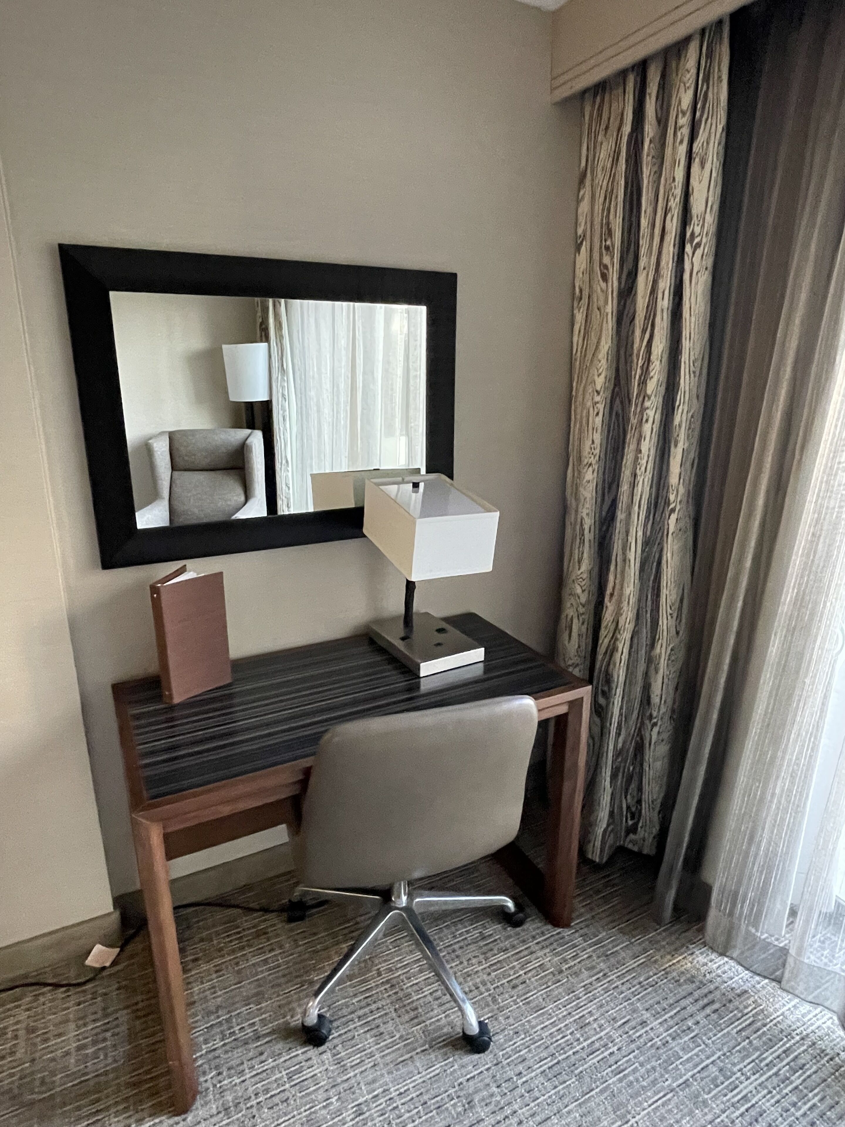 Habitación de hotel con un escritorio de madera, una silla, una lámpara, un espejo y una ventana con cortinas.