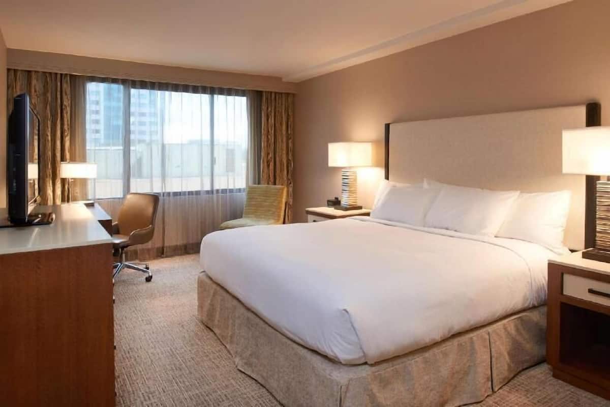 A hotel room with a large bed, a desk, a chair, two bedside tables with lamps, and a view of the city through large windows.