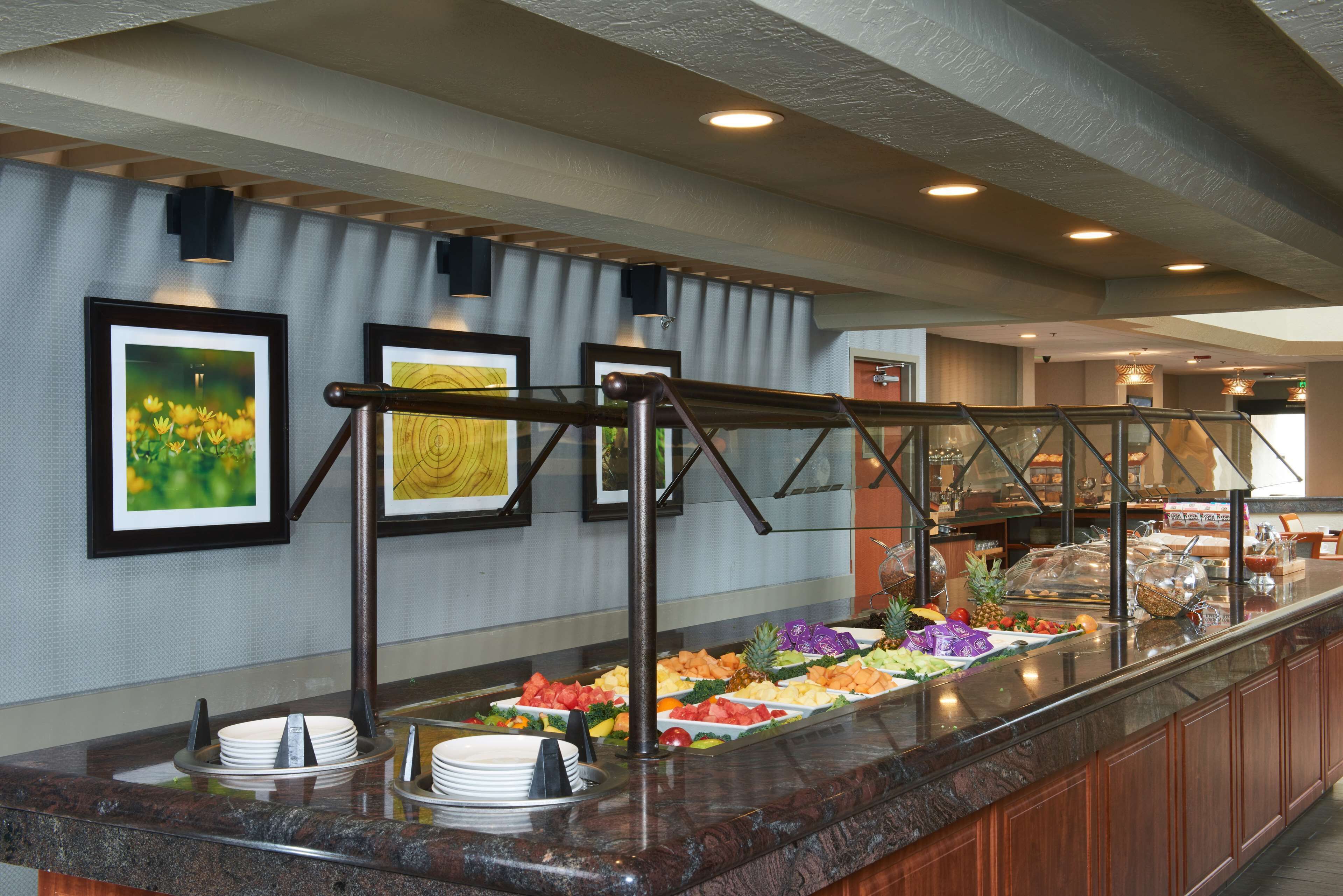 Zona de bufé con frutas y verduras variadas, barandilla de cristal y cuadros en la pared.
