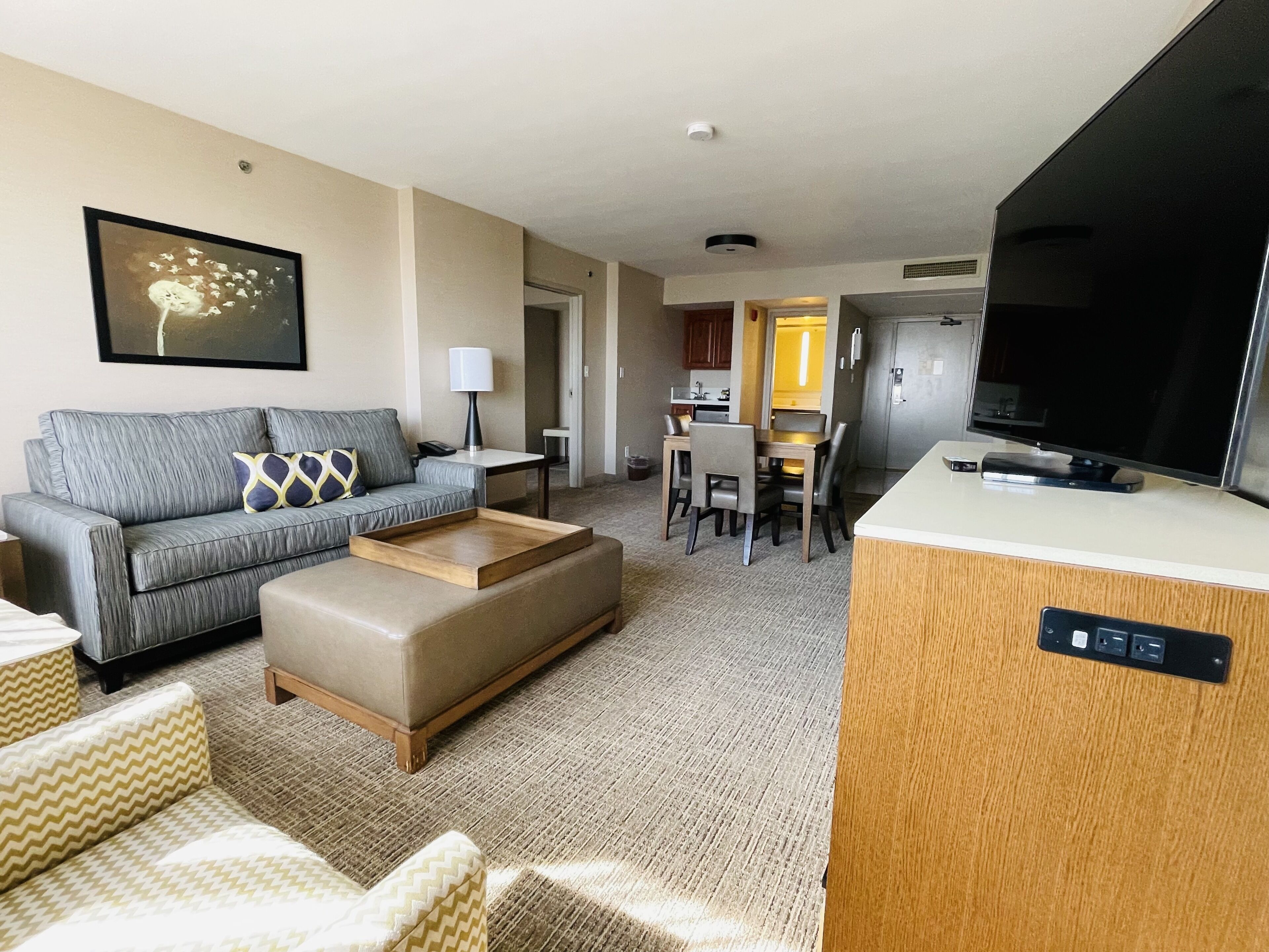 Une chambre d’hôtel avec un canapé gris, un pouf en bois, une table à manger avec des chaises, un téléviseur et un coin cuisine.