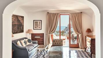 Una sala de estar acogedora con sofá, reposapiés y una mesita de centro. Cuenta con una chimenea, vistas al mar a través de un ventanal y un cuadro en la pared.