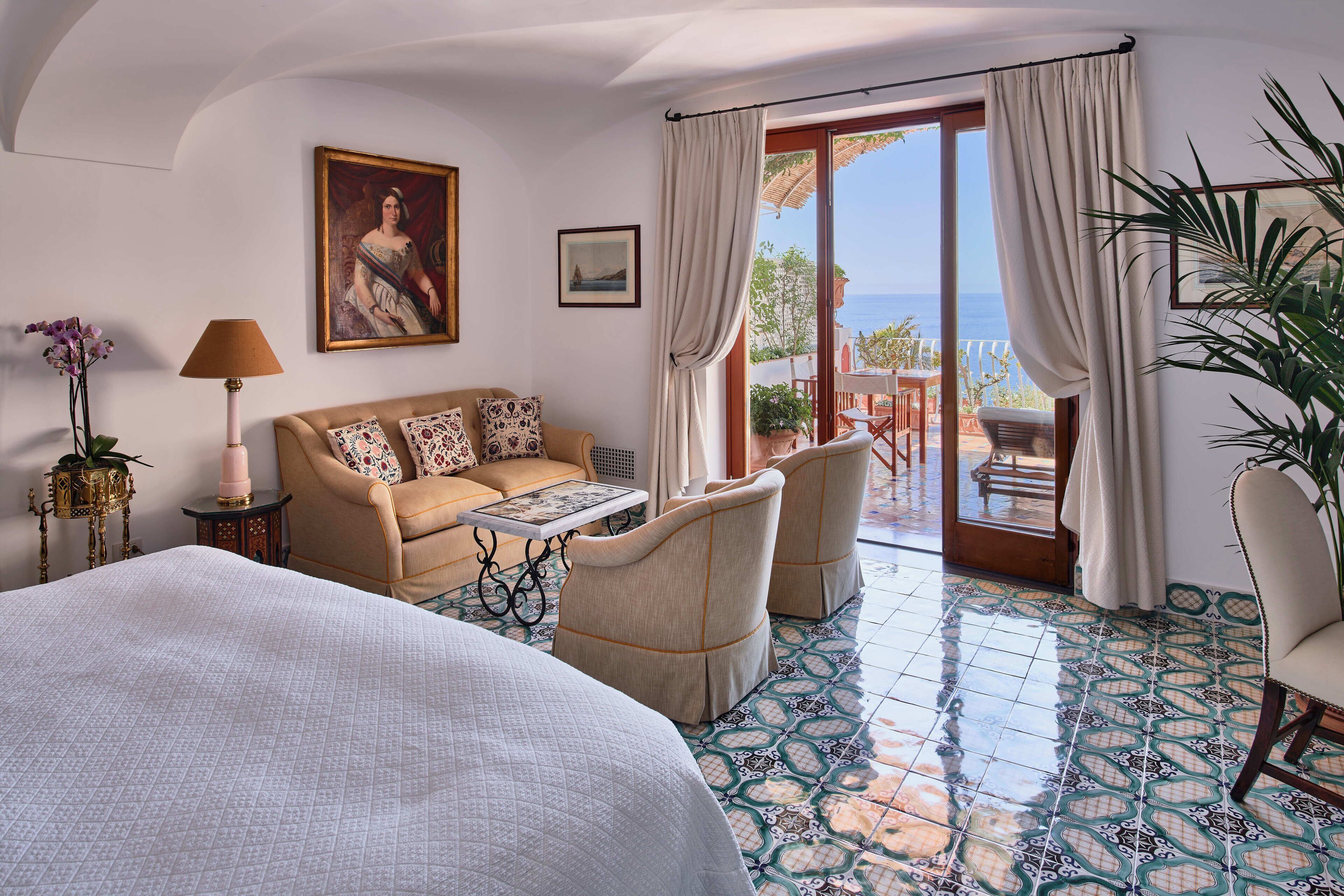 A hotel room with a bed, a sofa, a table, and chairs. There is a view of the sea through the glass doors.
