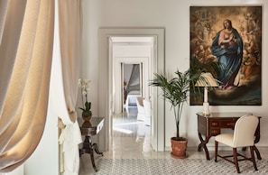 A hotel room with a bed, a desk, a chair, a potted plant, and a painting on the wall.