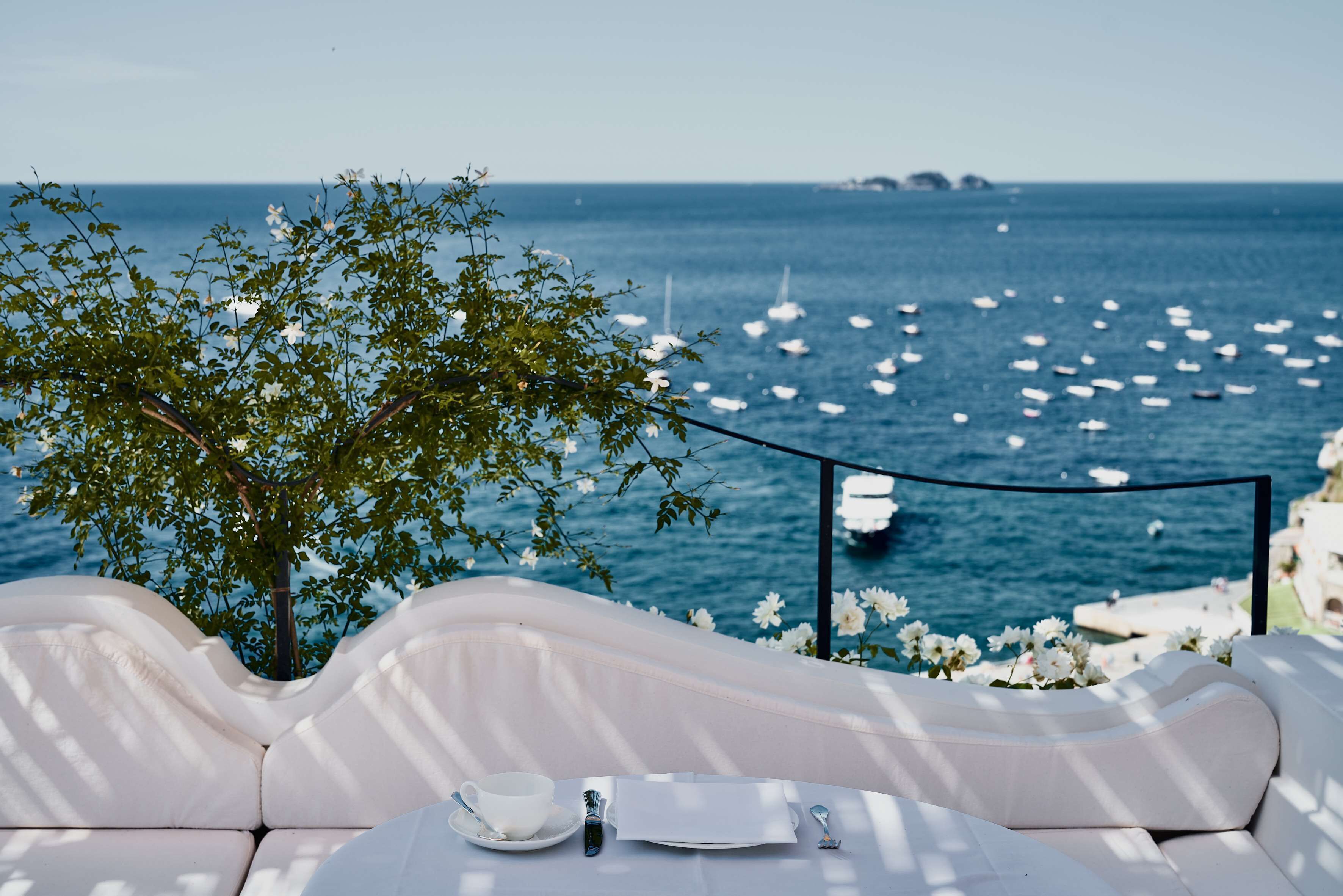Una terraza frente al mar con una zona de estar con cojines blancos, una mesa dispuesta con una taza, una cuchara y una servilleta, con vista a un mar tranquilo con barcos y un árbol con flores blancas.