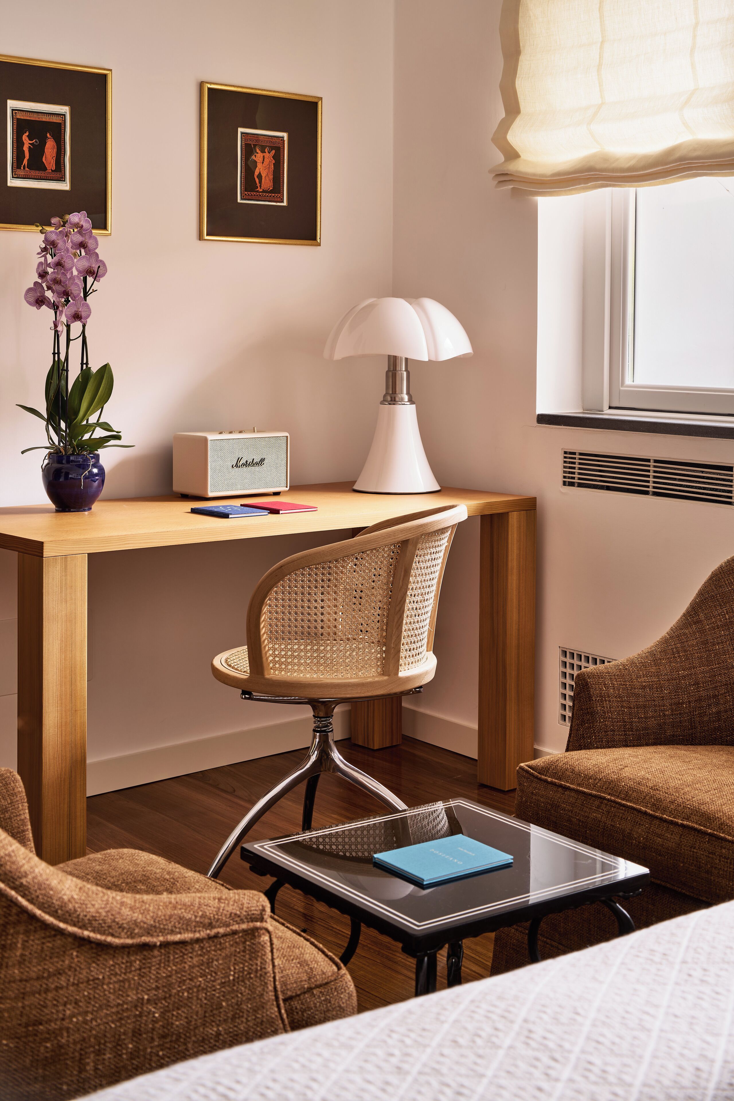 Ein Hotelzimmer mit einem Holztisch, einer weißen Lampe, einem Korbstuhl, einem braunen Sofa und einem Fenster mit Jalousien.