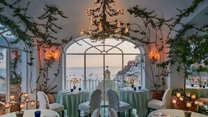A dining area with tables set for two, overlooking a scenic view of the sea and buildings. - Le Sirenuse (Positano)