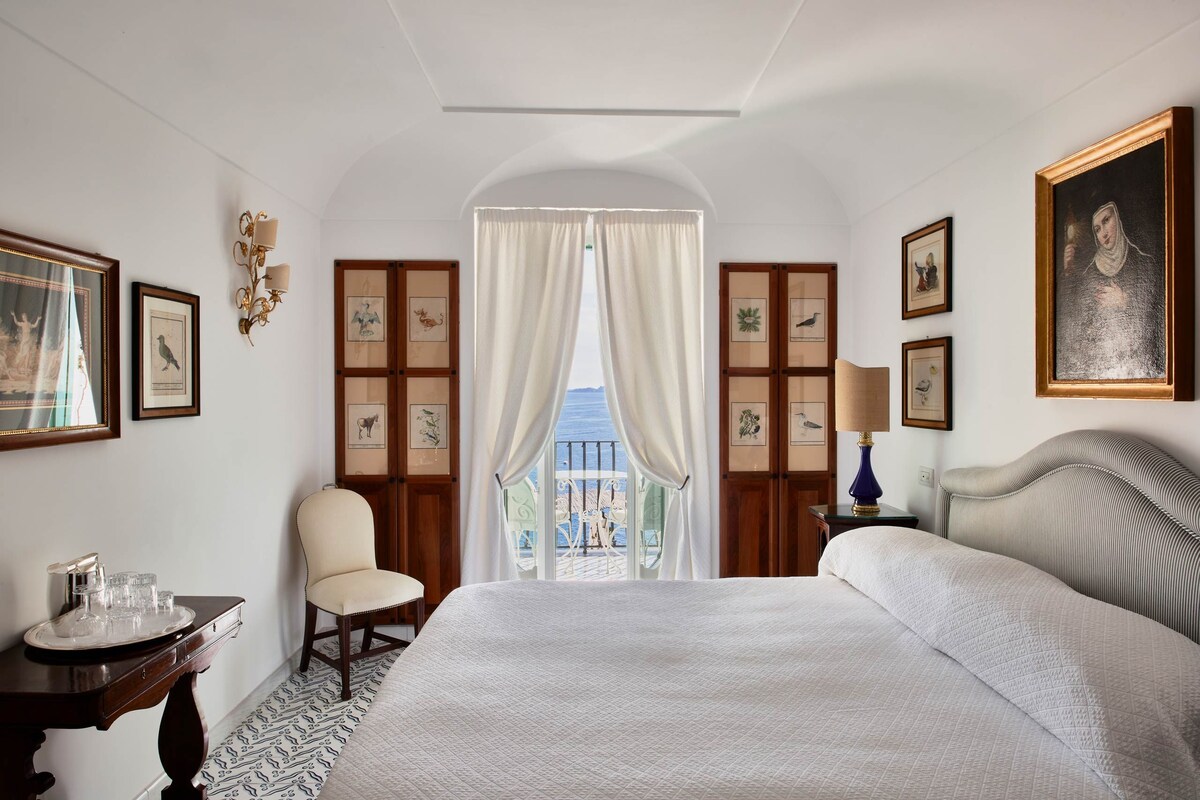 Une chambre d’hôtel avec un grand lit, une chaise, un bureau avec un plateau de verres et une vue sur le balcon.