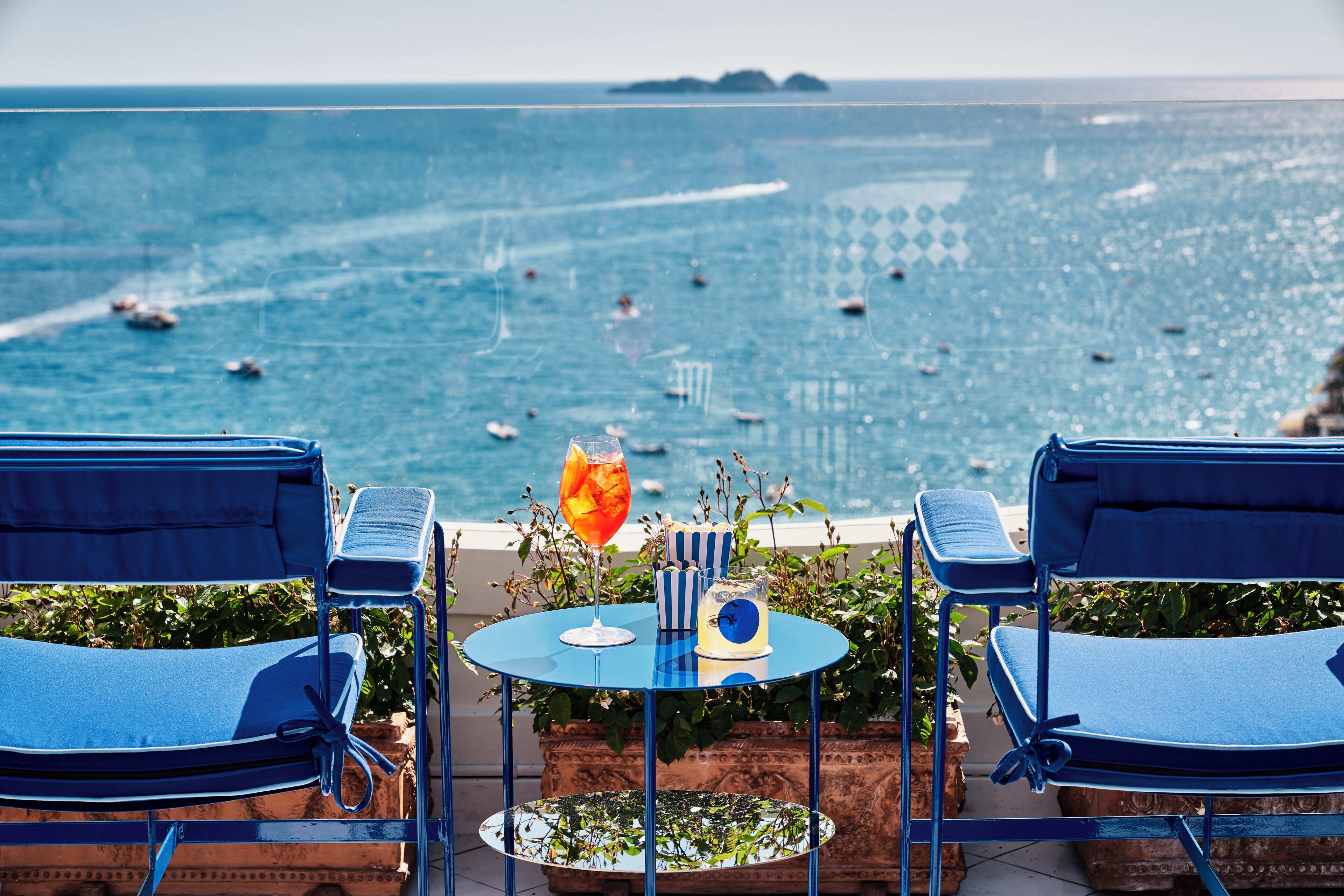 Una terrazza con due poltrone blu, un tavolino con una bevanda e vista sul mare con barche.