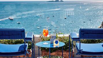 Una terrazza con due poltrone blu, un tavolino con una bevanda e vista sul mare con barche.