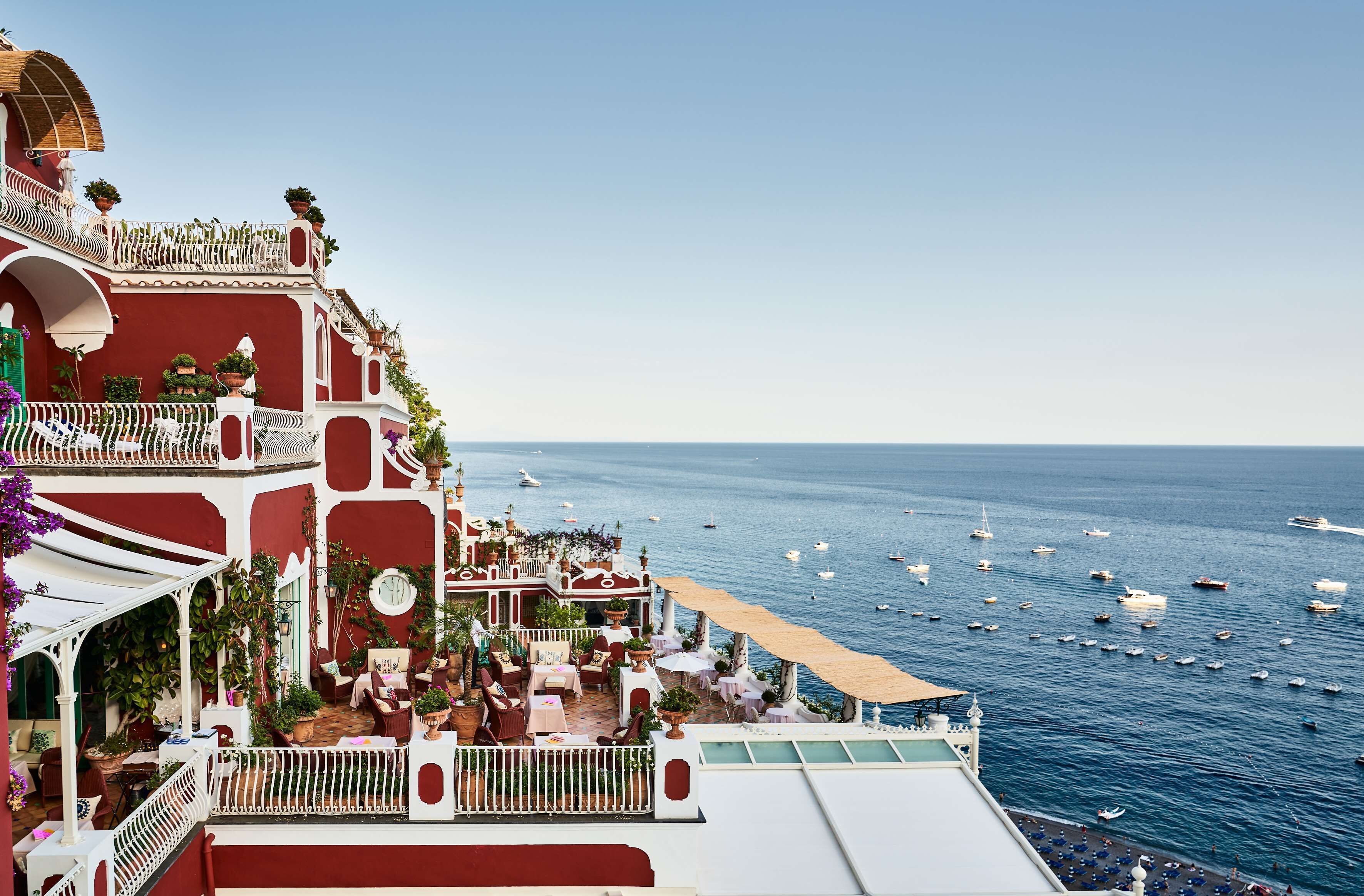 A coastal building with outdoor seating overlooking the sea, featuring balconies with potted plants and a view of boats.