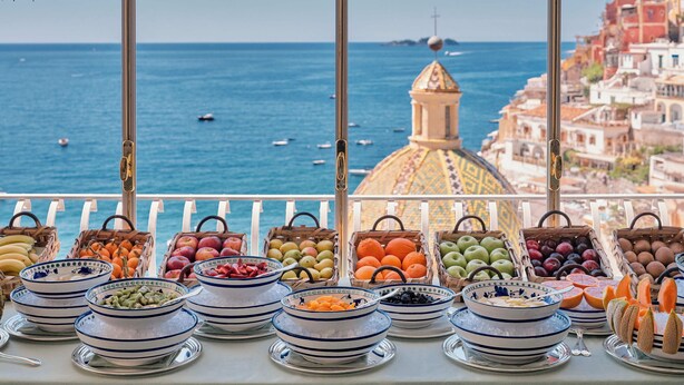 A buffet table with various fruits and vegetables, overlooking a coastal view with boats and buildings. - Le Sirenuse (Positano)