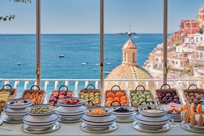 A buffet table with various fruits and vegetables, overlooking a coastal view with boats and buildings. - Le Sirenuse (Positano)