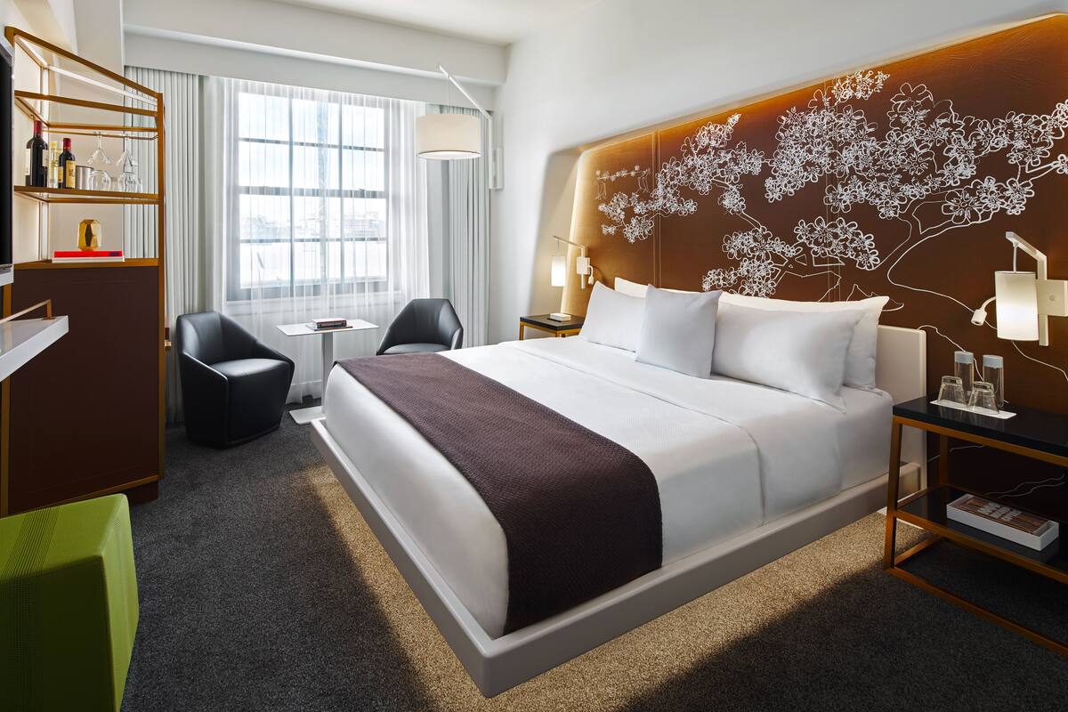 A modern hotel room with a large bed, a desk, a chair, and a bookshelf.