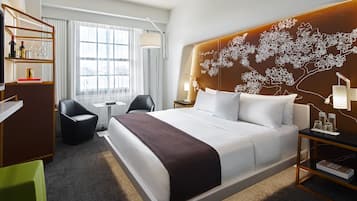 A modern hotel room with a large bed, a desk, a chair, and a bookshelf.