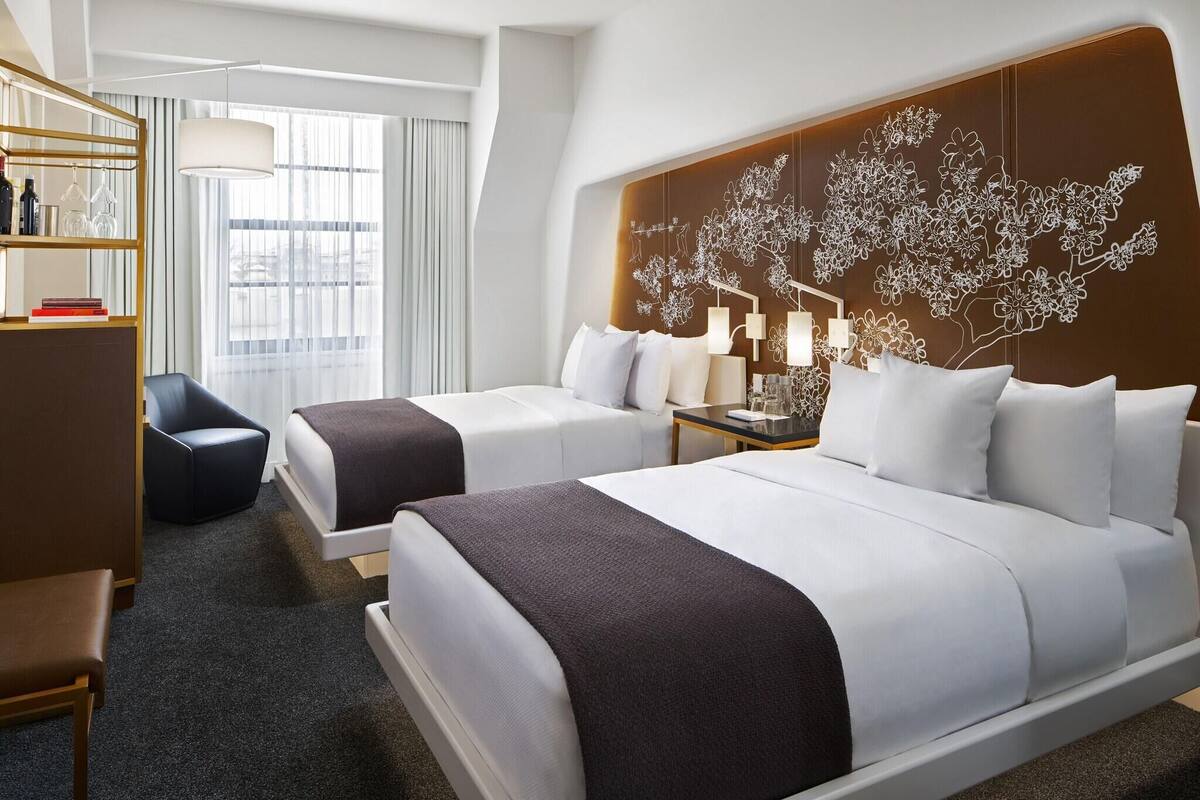 A hotel room with two beds, a desk, a chair, a bookshelf, and a large window.