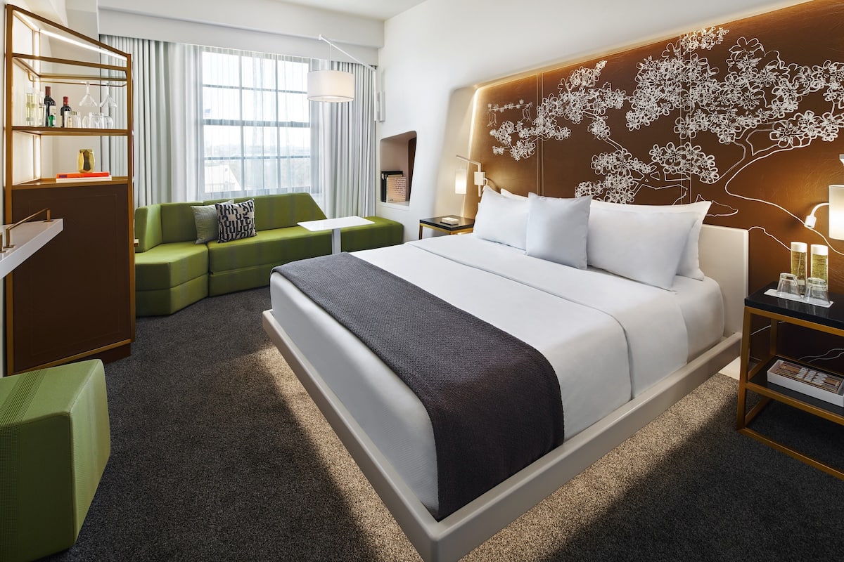 A modern hotel room with a large bed, a green sofa, a wooden headboard with a floral design, a small table with a lamp, and a bookshelf.