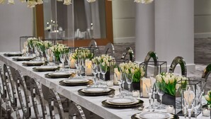 Une longue table à manger, dressée avec de l’argenterie blanche, des éléments décoratifs dorés, et entourée de fleurs blanches et de bougies.