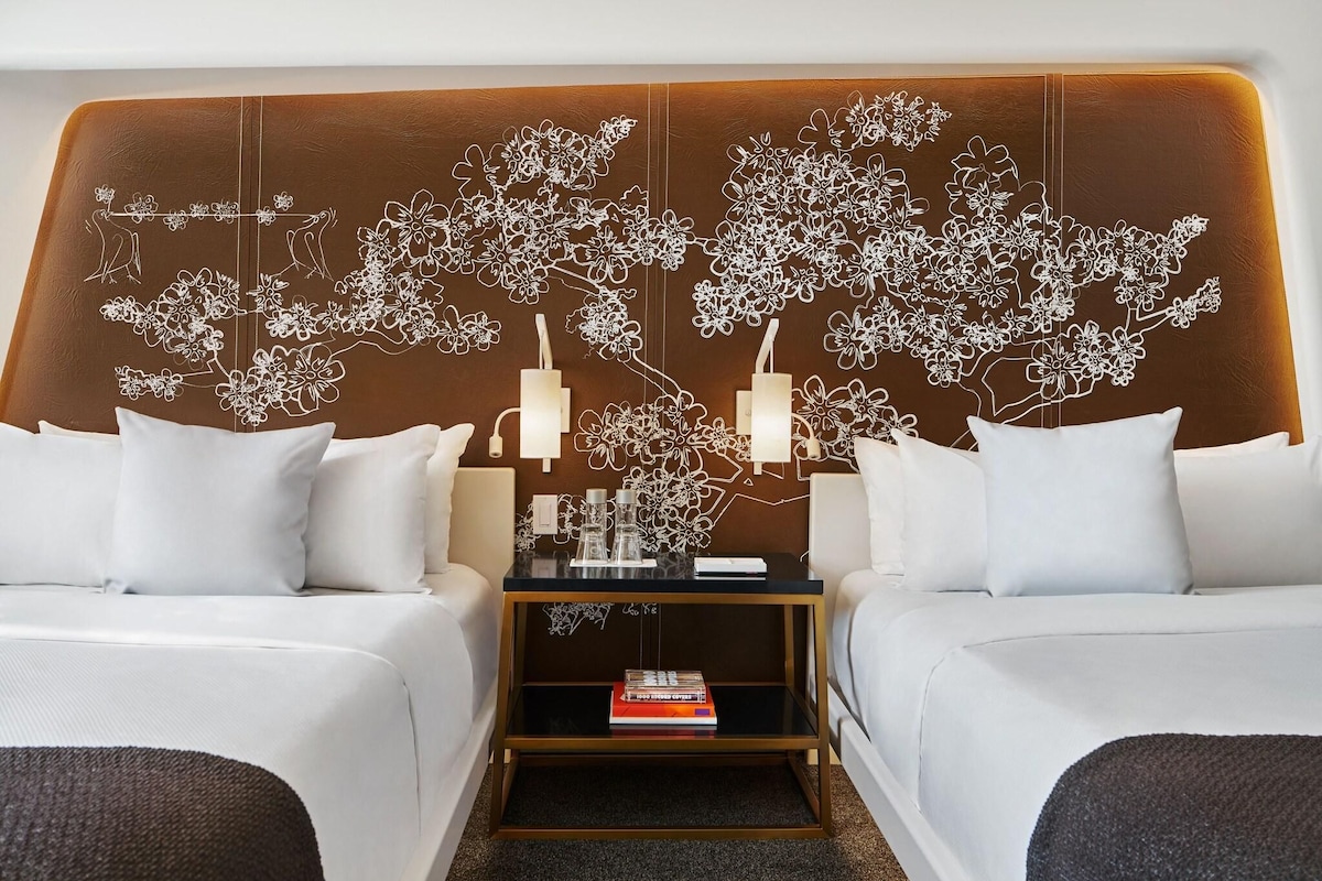 Una habitación de hotel con una cama grande, dos mesitas de noche con lámparas, un muro decorativo y un estante pequeño con libros.