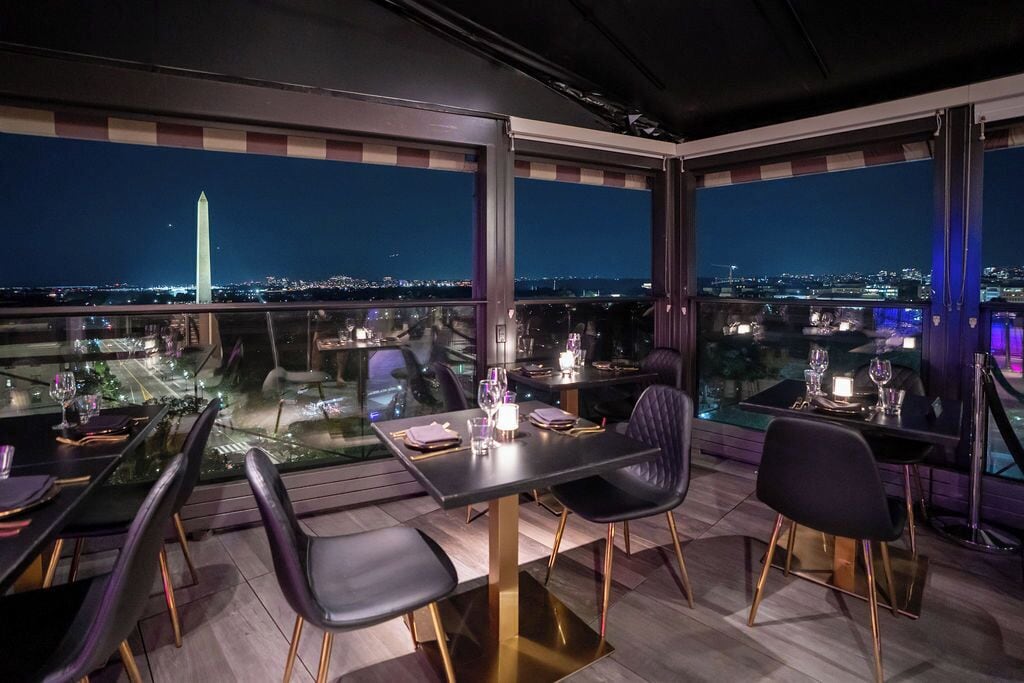 A rooftop dining area with tables and chairs, a city view, and the Washington Monument in the background.