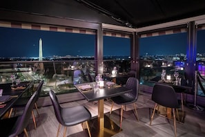 Un área de comedor en la azotea con mesas y sillas, vistas a la ciudad y el Monumento a Washington al fondo.