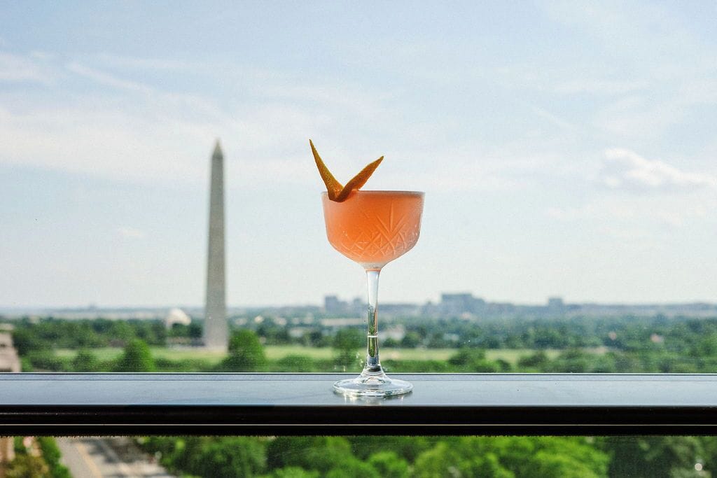 A cocktail glass with a garnish on a balcony overlooking a cityscape with a prominent monument.