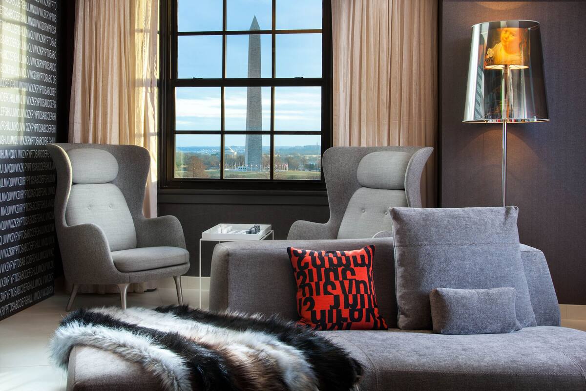 A modern living room with two grey armchairs, a grey sofa, a red and black patterned pillow, a fur throw, and a floor lamp.