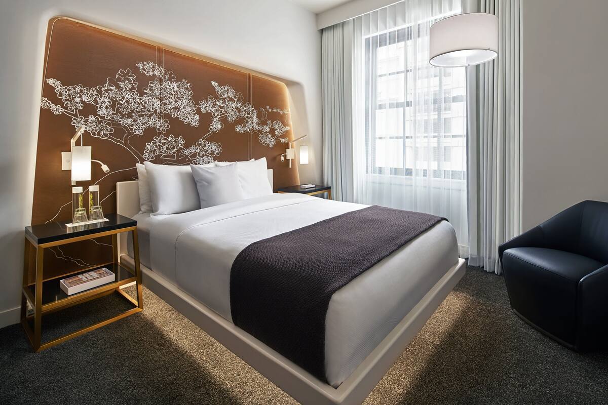 A modern hotel room with a large bed, a nightstand with a lamp, a reading area, and a view of the city through large windows.