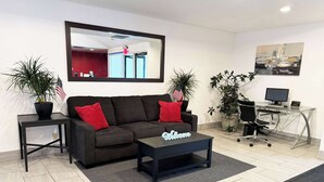 A modern living room with a dark brown sofa, two red cushions, a coffee table with a 'Welcome' sign, a desk with a computer, and two potted plants. -  Econo Lodge Wausau - Rothschild (Rothschild)