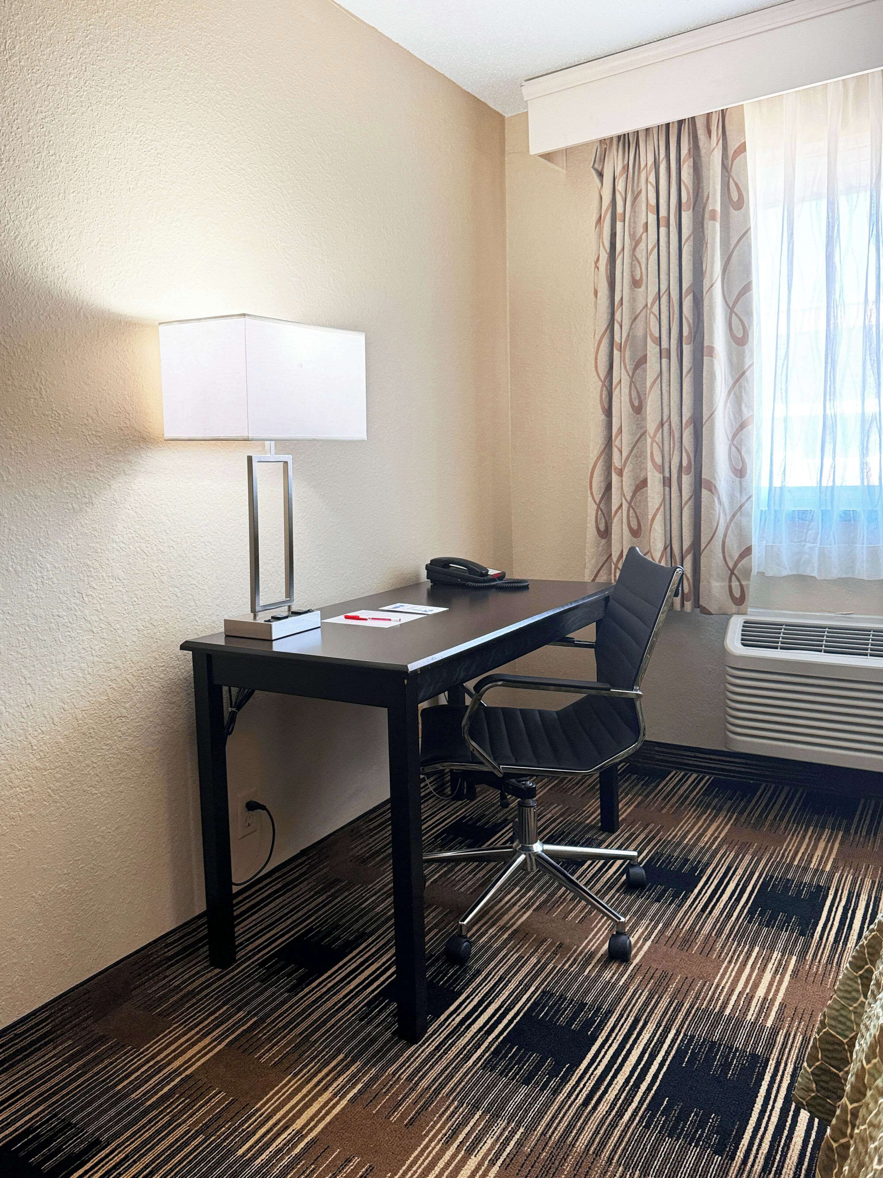 A hotel room with a desk, chair, lamp, phone, and air conditioning unit.
