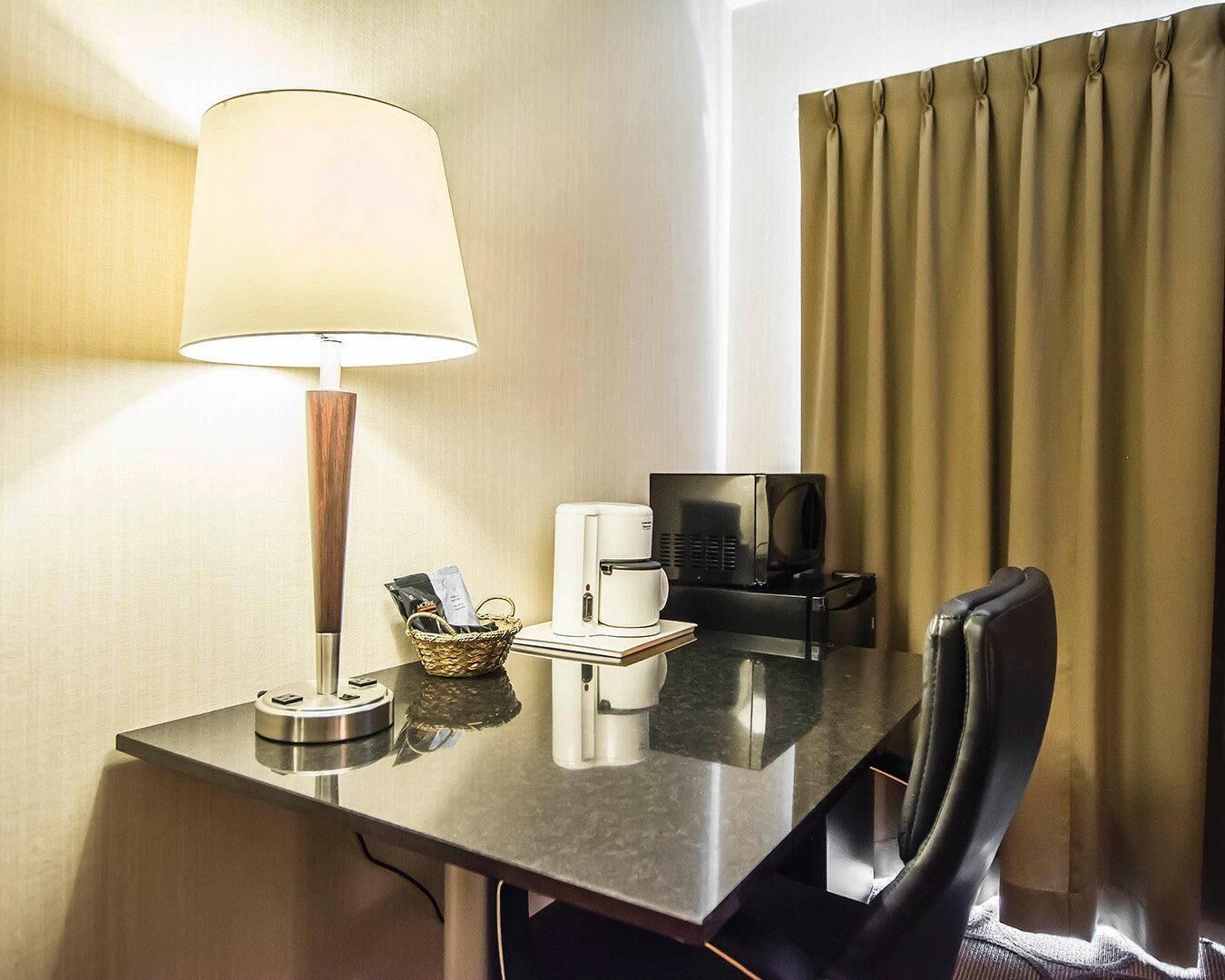 A hotel room with a desk, a lamp, a coffee maker, a TV, and a chair.
