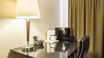 A hotel room with a desk, a lamp, a coffee maker, a TV, and a chair.