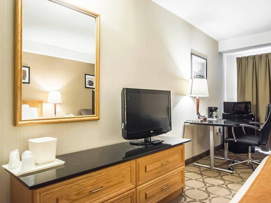 A hotel room with a flat-screen TV, wooden dresser, desk with a lamp, chair, and a large mirror.