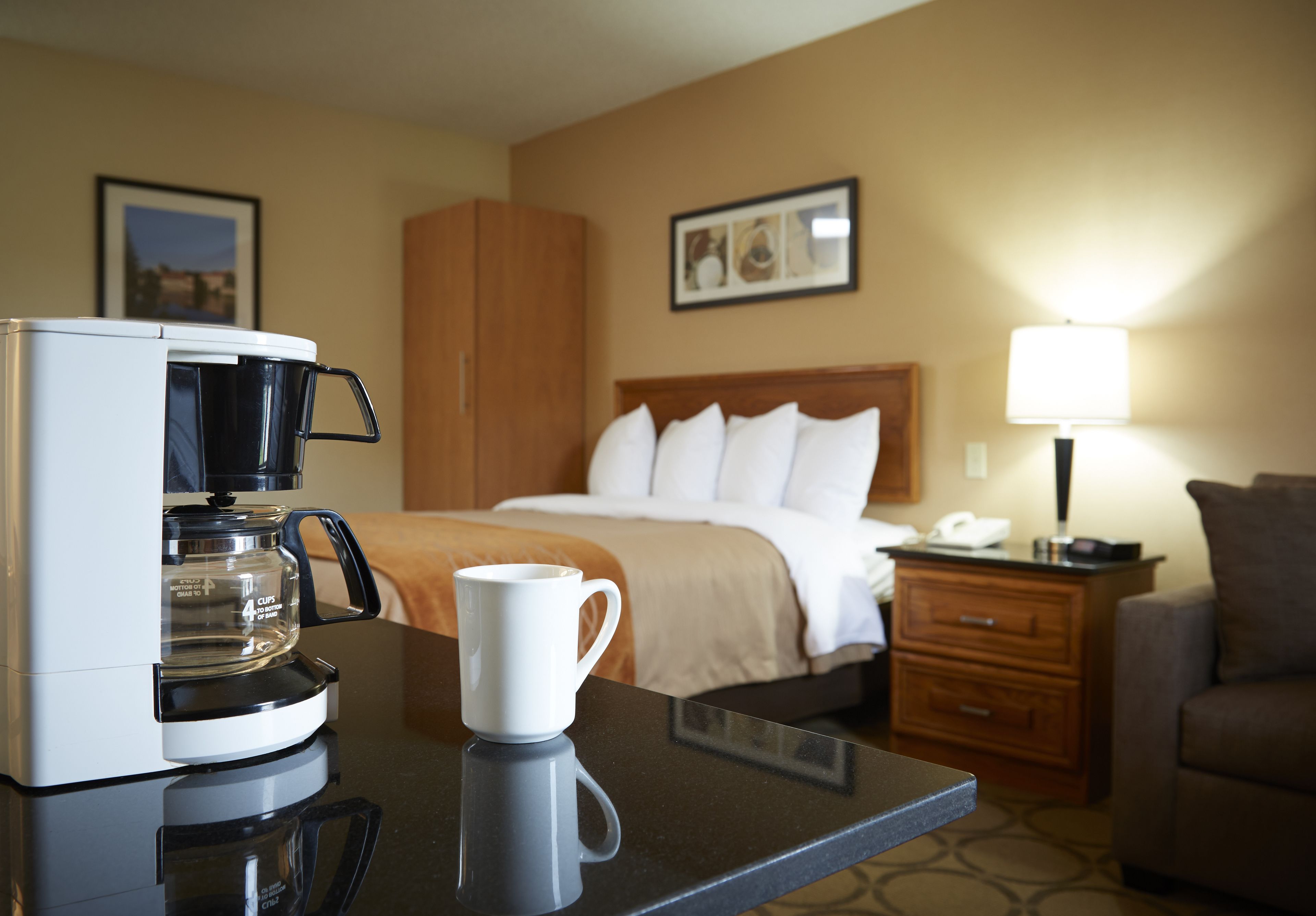 Habitación de hotel con una cama, una mesita de noche, una lámpara, una cafetera y una taza sobre una mesa.