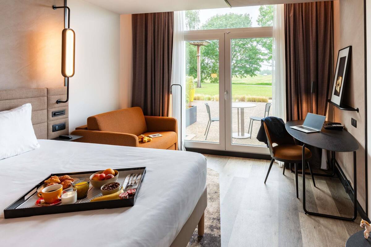 A hotel room with a bed, a desk with a laptop, a chair, a tray with breakfast, and a view of the outdoors.