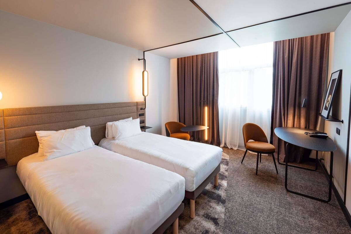 A hotel room with two beds, a desk, a chair, a TV, and a window with curtains.